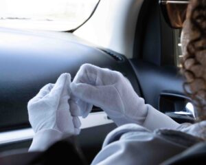 A backseat view of a woman with sunglasses and curly hair sitting in the passengers seat in a car, wearing white gloves.